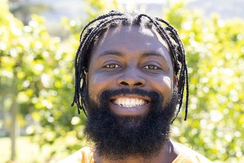 Smiling man with dreadlocks and full beard wearing orange shirt in sunlit park