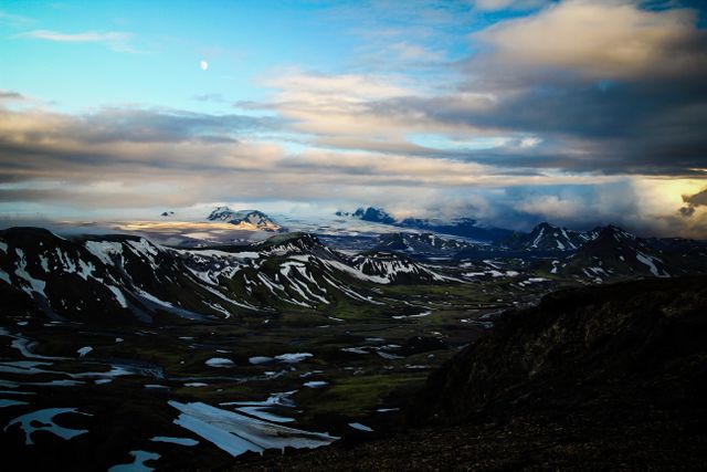 Snow-capped mountains with a twilight backdrop, partly cloudy sky, and scattered snow patches create a dramatic and serene landscape. Ideal for use in travel brochures, nature-themed websites, and environmental campaigns, this scene captures the raw beauty and tranquility of remote wilderness.