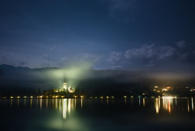 Night scene of Church on Lake Bled with its reflection seen in calm water. Sky is clear with a few stars visible, enhancing tranquil and peaceful atmosphere. Ideal for travel blogs, relaxation themes, or nature-related content.