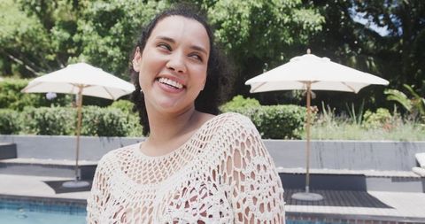 Smiling woman wearing white crocheted cover-up standing by sunny pool with umbrellas