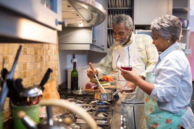 Senior couple cooking together in modern kitchen, smiling and enjoying each other's company. Man stirs pot on the stovetop while woman holds wine glass, contributing to meal preparation. Suitable for topics related to family life, domestic happiness, elderly lifestyle, cooking hobbies, and culinary activities at home.