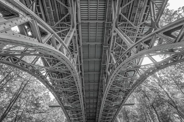 Detailed view of the underside of an iron bridge captured in black and white. The image showcases the intricate architectural and engineering elements with symmetrical lines and metal framework. Ideal for use in articles, presentations, or creative projects related to engineering, architecture, urban infrastructure, or structural design.