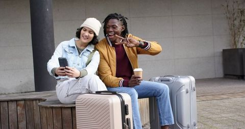 Young diverse travelers sitting with luggage and smartphone at urban plaza