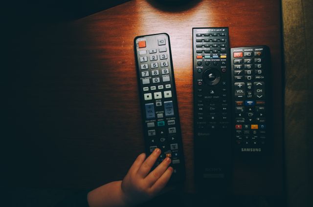 A child hand is picking up a remote control from a collection placed on a wooden table. This could be used to represent concepts of technology interaction, family life, and controlling media devices in a home environment. Suitable for technology blogs, electronic product advertisements, and family-oriented publications.