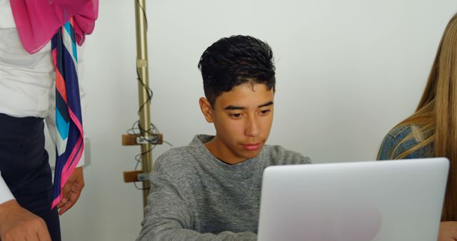 Young male student concentrating on laptop task in classroom setting, with educator providing assistance. Ideal for use in educational content, technology in education themes, tutoring services, study aids, and student success stories.