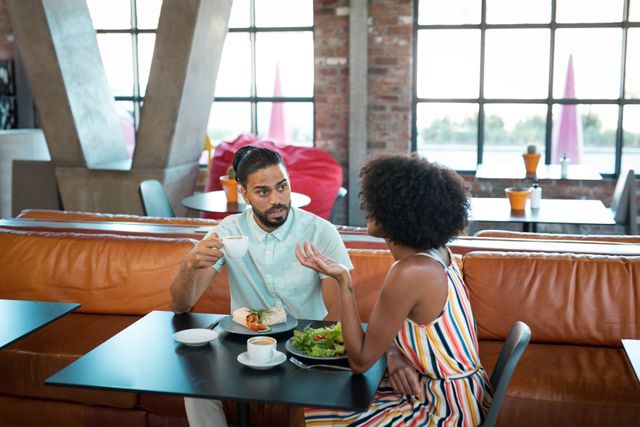 Diverse couple enjoying lunch and coffee in a modern cafe, engaging in conversation while looking at a smartphone. Ideal for use in lifestyle blogs, social media posts, advertisements for cafes or restaurants, and articles about relationships and communication.
