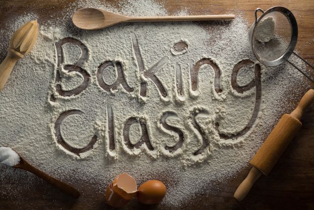 This image shows the words 'Baking Class' written in flour on a wooden surface, surrounded by various baking tools such as a wooden spoon, rolling pin, sieve, and eggshells. Ideal for promoting baking courses, culinary workshops, cooking blogs, or recipe websites.