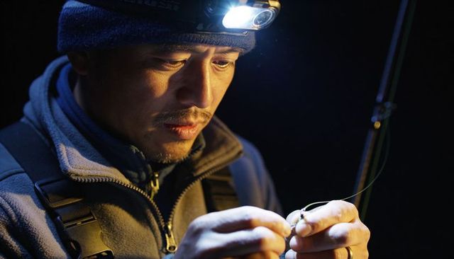 Night angler tying hook with headlamp, preparing rod for riverbank fishing