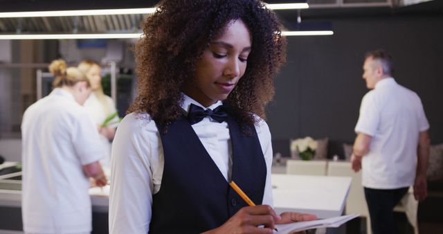 Biracial woman wearing vest and bow tie writing in notebook in kitchen. Lifestyle, learning, food, cooking, unaltered.