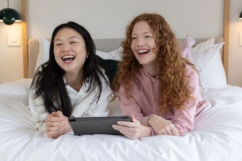 Diverse female friends laughing and watching tablet while lounging on bed with white duvet