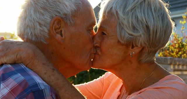 A senior Caucasian couple shares a tender kiss in a warm, sunlit setting, with copy space. Their affectionate moment captures a timeless expression of love and companionship.