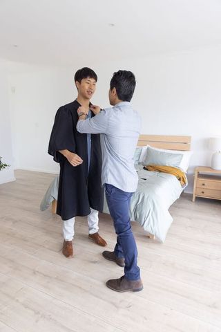 Asian graduate getting graduation gown adjusted by friend in bright minimalist bedroom