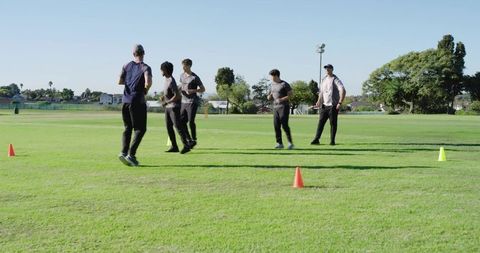 Multiracial men performing high-knee warm-up drill on green field with coach guiding team