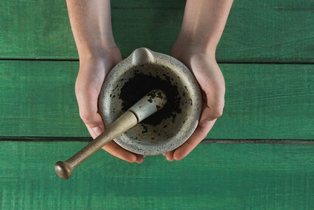Hands holding a mortar and pestle with herb paste on a green wooden table. Ideal for use in articles or advertisements related to traditional medicine, herbal remedies, organic ingredients, and natural health practices. Perfect for illustrating concepts of alternative medicine, herbal preparation, and rustic kitchen tools.
