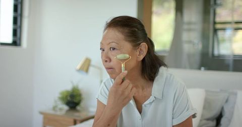 Senior asian woman applying jade roller on cheeks in minimalist bedroom skincare routine