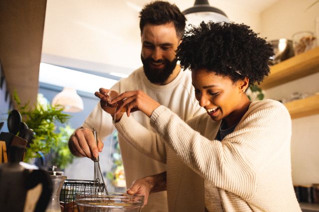 Happy biracial young woman breaking egg and boyfriend mixing batter in bowl in kitchen. Unaltered, lifestyle, love, togetherness, cooking, food, afro hair, beard and home concept.