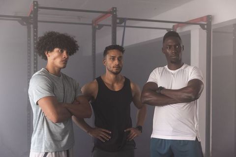 Three diverse men standing with arms crossed watching squat rack demonstration in gym