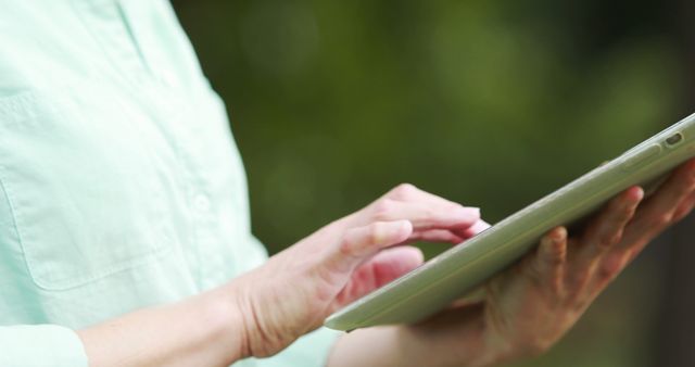 Person is using a tablet outdoors with greenery in the background, emphasizing technology use in natural settings. This image can be used for articles, blogs, or advertisements focused on technology, mobile devices, connectivity, or relaxation outdoors.