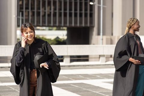 Graduates celebrating on urban campus plaza, holding mortarboards and talking on phone