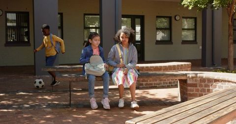 Diverse girls sitting on bench holding backpacks near soccer ball in school courtyard