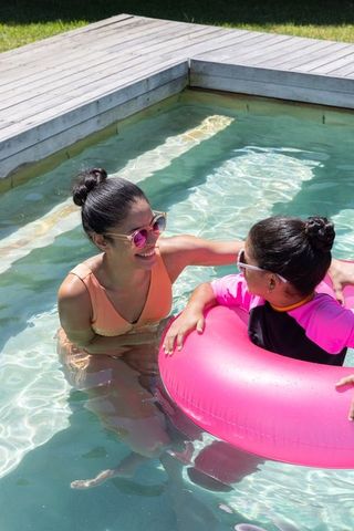 African american mother and daughter playing in backyard pool with pink float and sunglasses
