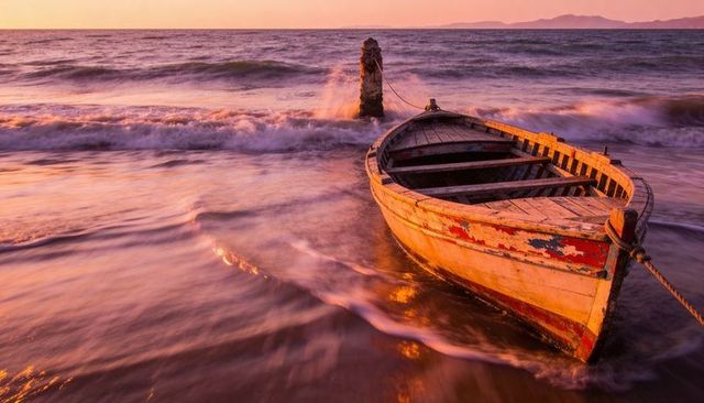 Weathered wooden rowboat bobbing in golden sunset surf holding to mooring post