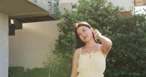 Young woman in pale yellow sundress touching hair in sunlit residential garden