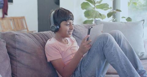 Preteen boy wearing headphones using tablet while relaxing on modern living room couch