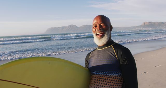 A senior man standing on a beach at sunrise, smiling and holding a yellow surfboard. The ocean waves and distant mountains form the background. This image is great for use in promotions for active living, surfing events, wellness campaigns, or retirement lifestyle content.