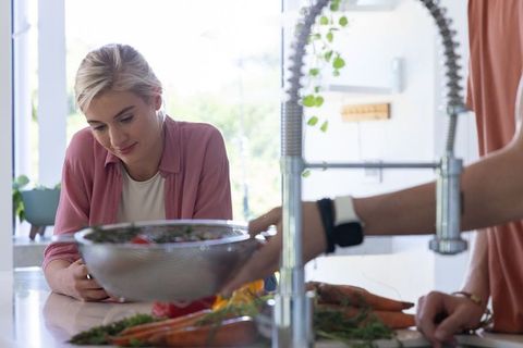 Couple rinsing fresh salad in modern sunlit kitchen, woman leaning on counter watching