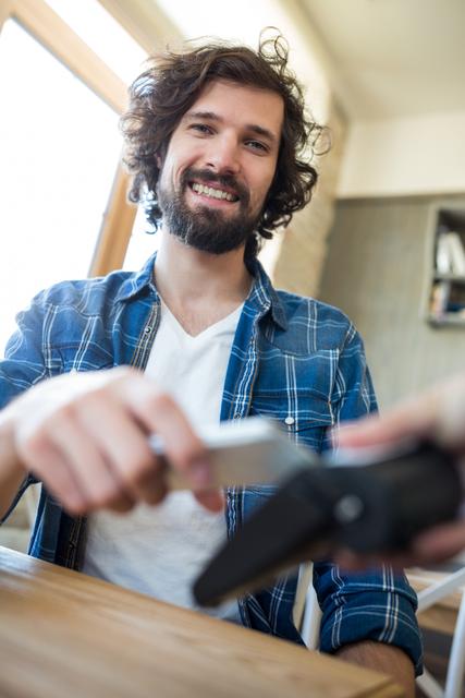 Young man with beard smiling while paying with NFC technology on mobile phone in coffee shop. Perfect for promoting modern payment solutions, contactless transactions, and casual lifestyle themes.