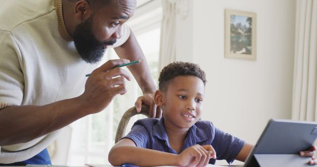 Father assisting his son with schoolwork using a digital tablet, capturing a moment of educational bonding and modern learning at home. This scene showcases positive parent-child interaction and the use of technology for educational purposes, perfect for use in articles related to parenting, education, family, and technology in learning.