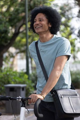 Smiling african man standing with bicycle in park wearing blue tee and crossbody satchel