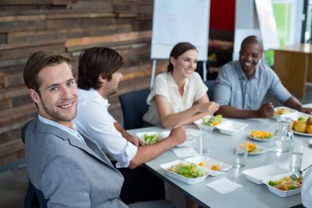 Business executives sitting around a table enjoying a meal together in an office environment. They are smiling and appear to be engaged in a casual discussion, fostering teamwork and collaboration. Ideal for use in corporate websites, business presentations, and promotional materials highlighting a positive work culture and team bonding.