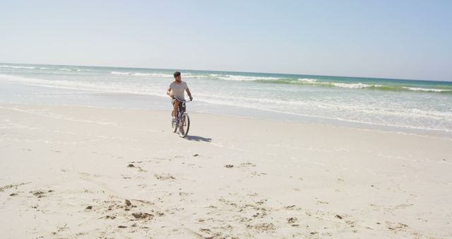 A young Caucasian man rides a bicycle along the shore on a sunny beach day, with copy space. His leisurely activity suggests a relaxed and enjoyable moment in a serene coastal environment.
