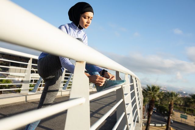 Biracial woman wearing hijab and sportswear tying her shoelaces outdoors in a city environment on a sunny day. Ideal for promoting urban fitness, active lifestyle, women's health, and inclusive sportswear. Suitable for use in fitness blogs, health and wellness articles, and advertisements for sports apparel.