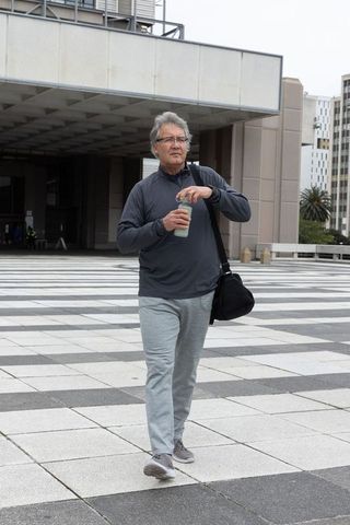 Senior asian man walking across modern plaza carrying black bag and water bottle