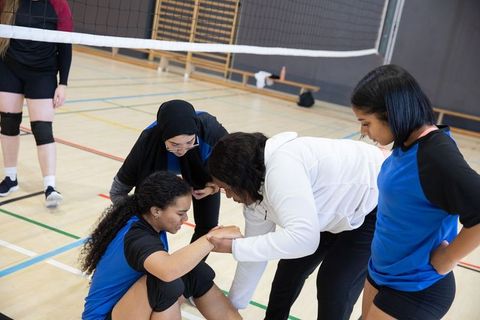 Coach helping injured teen volleyball player while teammates offering support on gym court