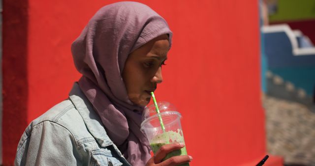 A young woman wearing a hijab and denim jacket is drinking a matcha green tea beverage while using her smartphone. The bright pink wall and other colorful buildings in the background add vibrant contrasts, making it a lively scene. This photo can be used for content related to youth lifestyle, modern technology use, casual fashion, or cultural representation in public spaces.