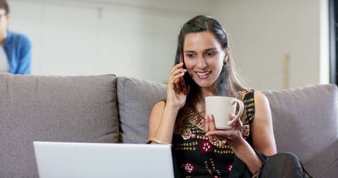 Indian woman working remote on sofa, talking on phone, holding mug and checking laptop