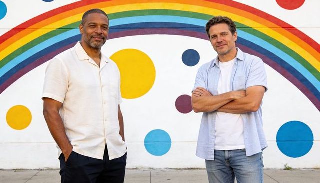 Two friends standing confidently in front of colorful rainbow mural on urban sidewalk