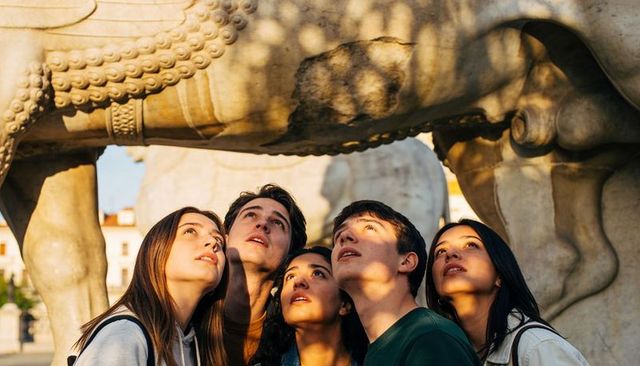 Teens looking up beneath historic stone sculpture in urban plaza at golden hour