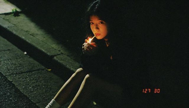 Asian teen lighting cigarette on curb at night with lighter glow and vintage red timestamp