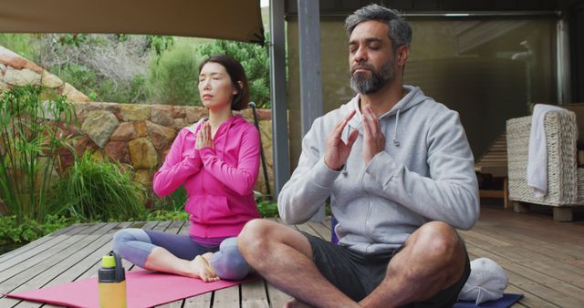 Happy diverse couple doing yoga, sitting and meditating at terrace. Spending quality time at home.