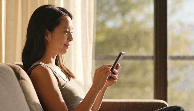 Asian woman relaxing on sofa using smartphone by sunlit window