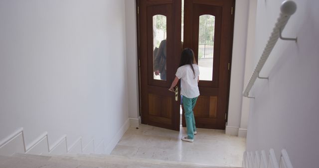 Child in casual clothing opening a wooden front door from an inside staircase of a home. Natural light is streaming through polished door panels, creating a welcoming atmosphere. Suitable for concepts of family, home living, childhood, security, and everyday moments.