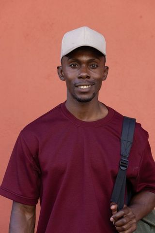 African man smiling in burgundy tee and beige cap holding backpack strap by terracotta wall