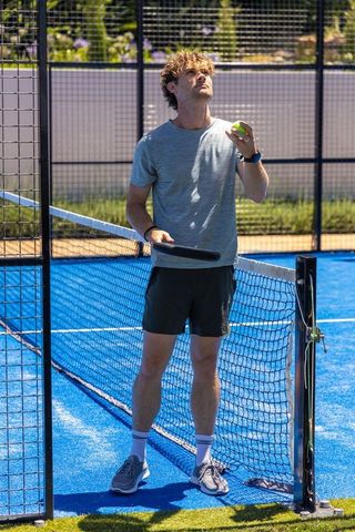 Young man standing at net on bright blue paddle court holding paddle and yellow ball