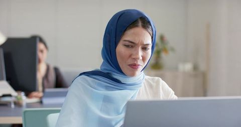 Indian muslim woman wearing blue hijab frowning at laptop in modern office environment