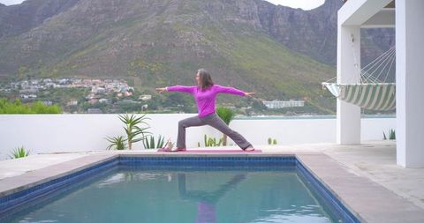 Mature woman practicing yoga by reflective pool on terrace with mountain view and hammock
